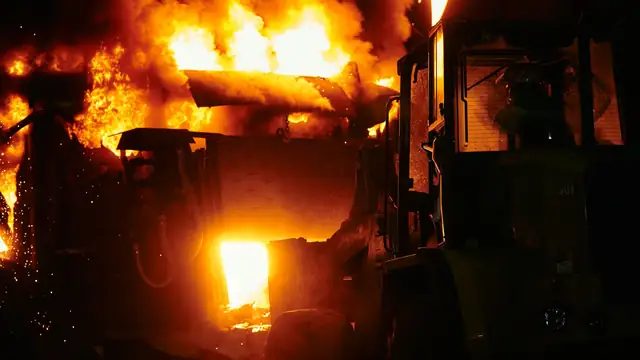Incêndio industrial à noite, com chamas e faíscas iluminando máquinas pesadas