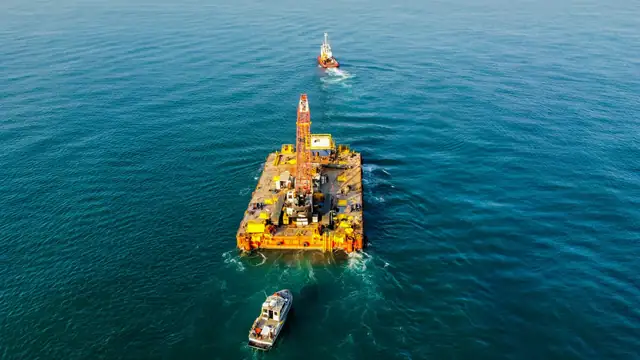 Vista aérea de equipamento pesado sobre uma balsa no mar, apoiada por rebocadores