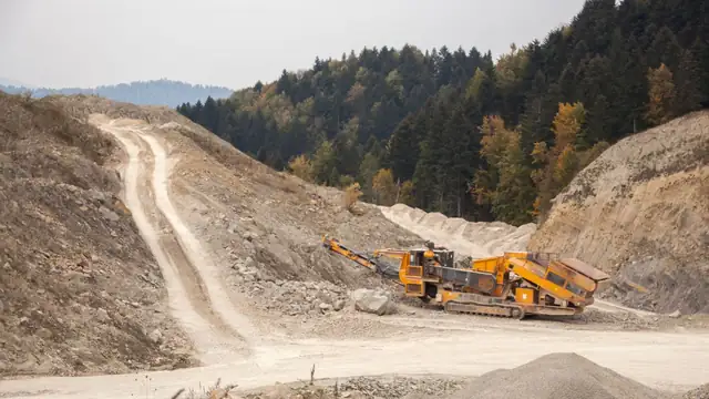Máquina de britagem industrial ao lado de grande pilha de terra, cercada por árvores