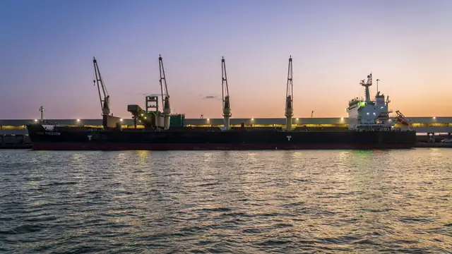 Navio graneleiro atracado em porto com guindastes ao pôr do sol, refletido na água