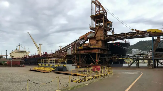 Porto industrial movimentado, com navios, guindastes e cais de carregamento sob céu nublado