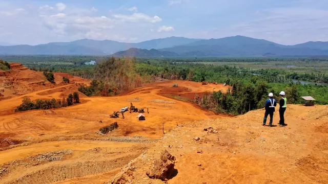 Duas pessoas observam área de mineração em solo vermelho, com morros ao fundo